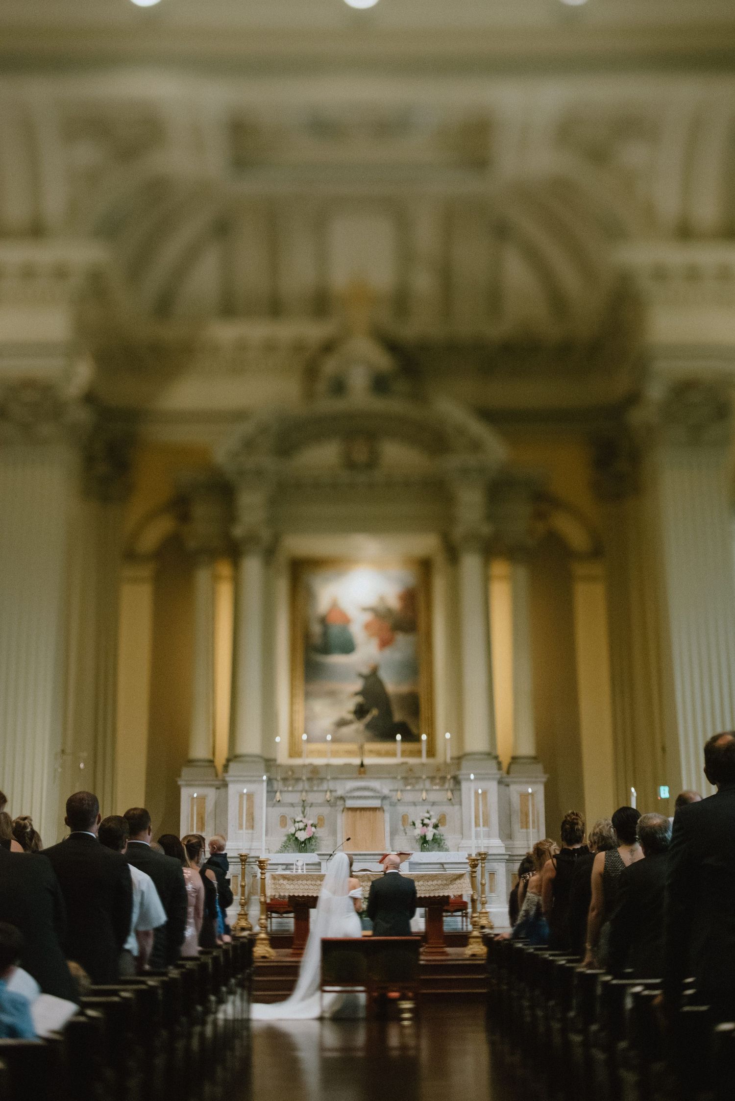 Dramatic view down church aisle during wedding ceremony with ornate altar in soft focus.