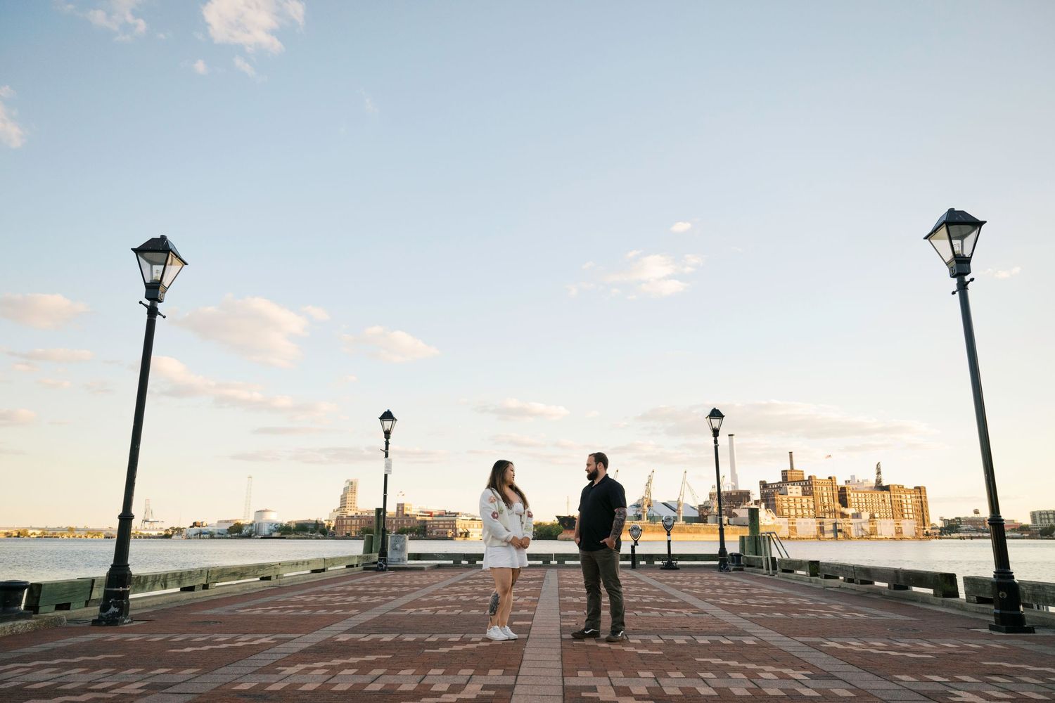 Couple stands on brick boardwalk at sunset with vintage lamp posts and harbor view in Baltimore.