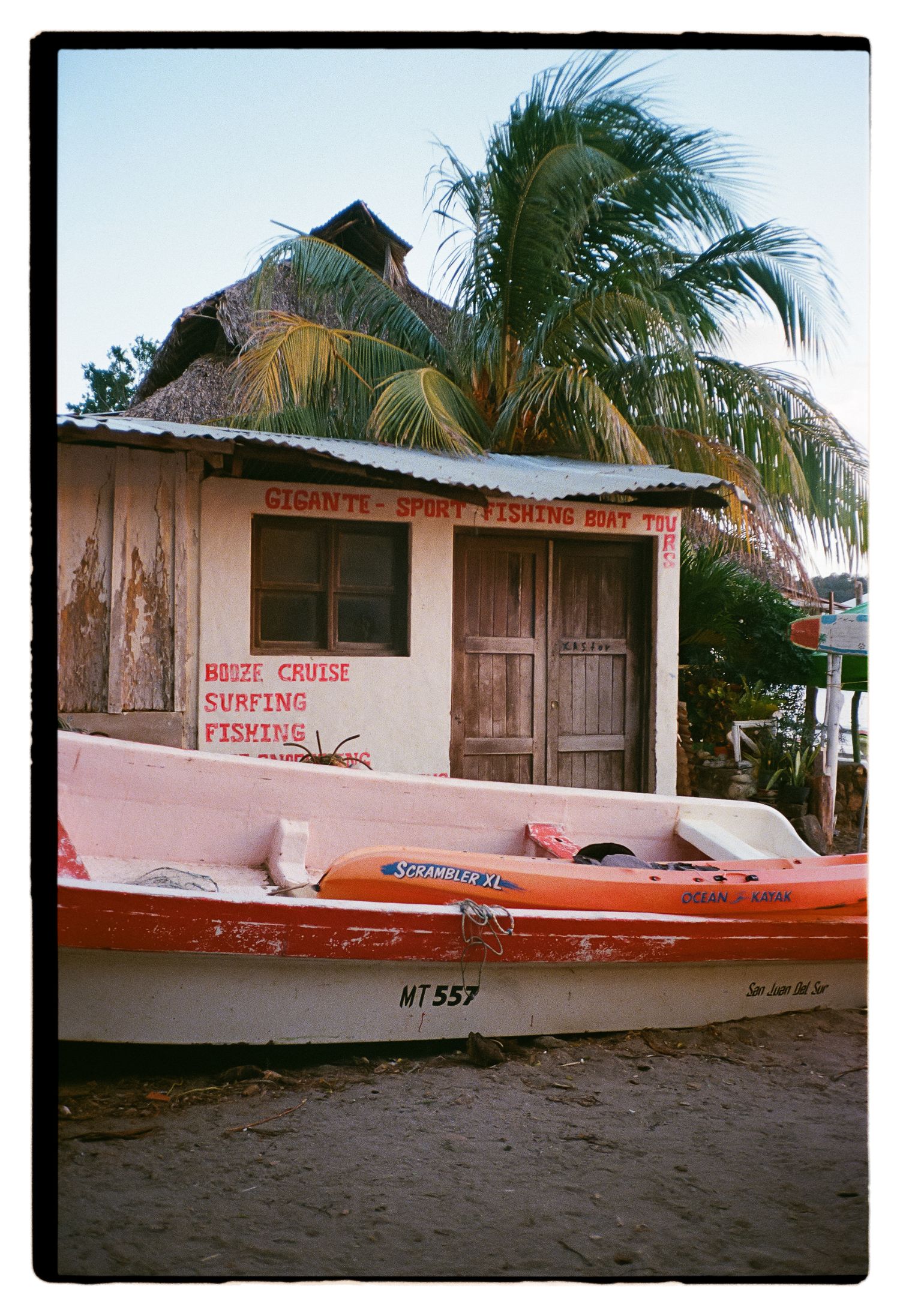 A vintage fishing shack with palm trees and a red boat parked in front on a sandy beach.