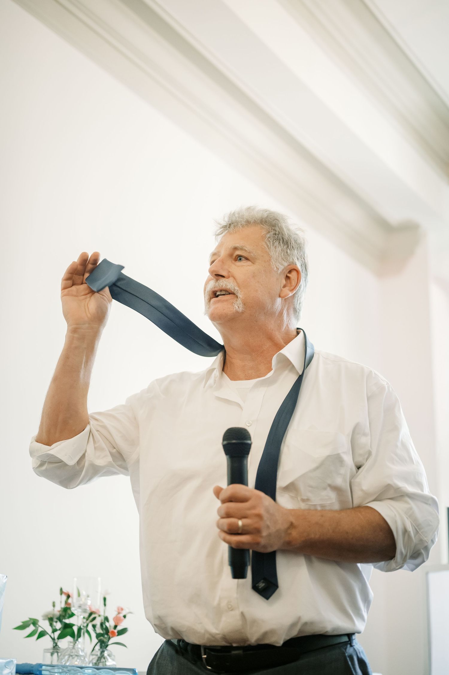 Speaker holds up tie while giving speech at wedding reception in bright indoor venue.