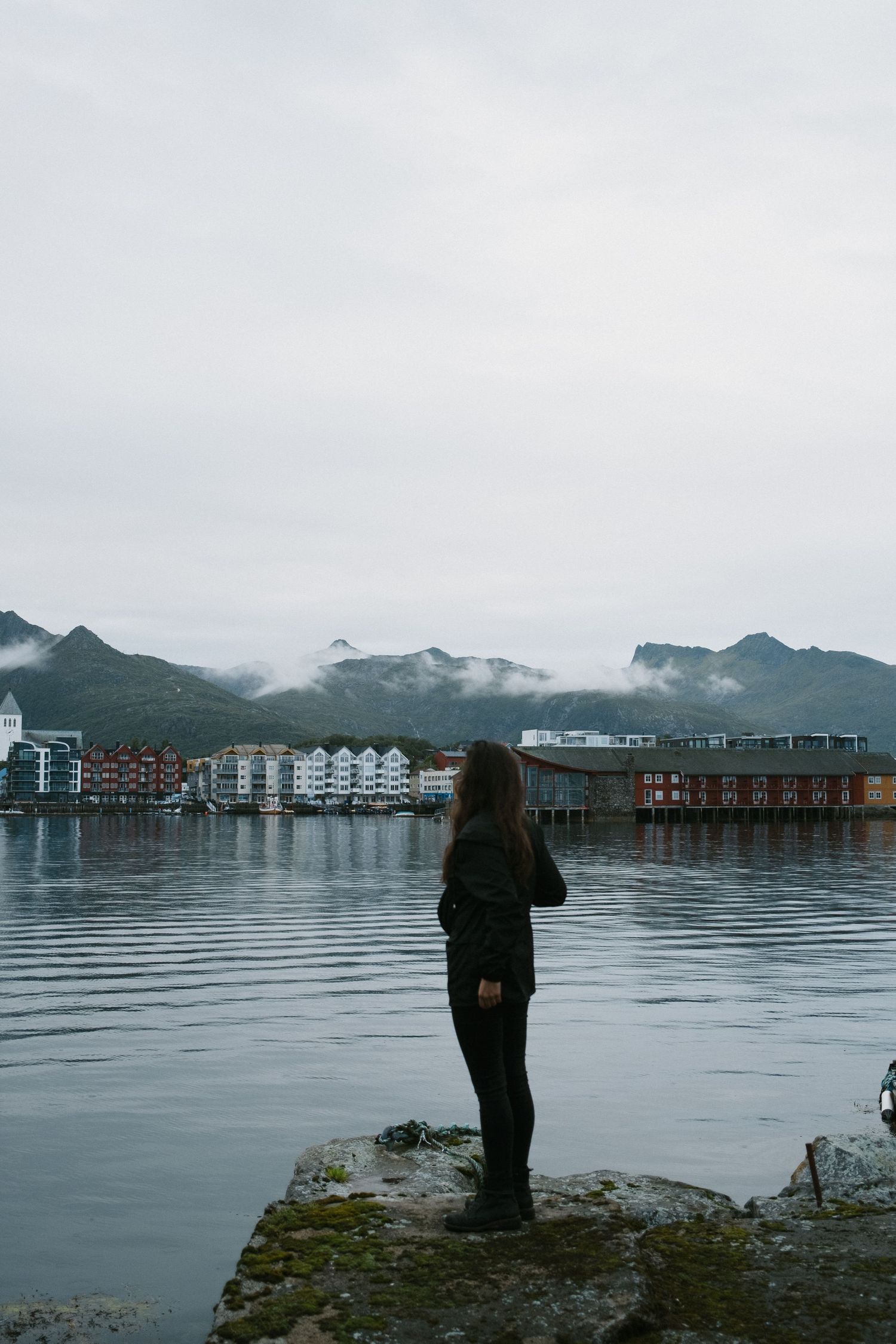 Silhouetted figure stands on rocky shore overlooking waterfront buildings and misty mountains in Norway.