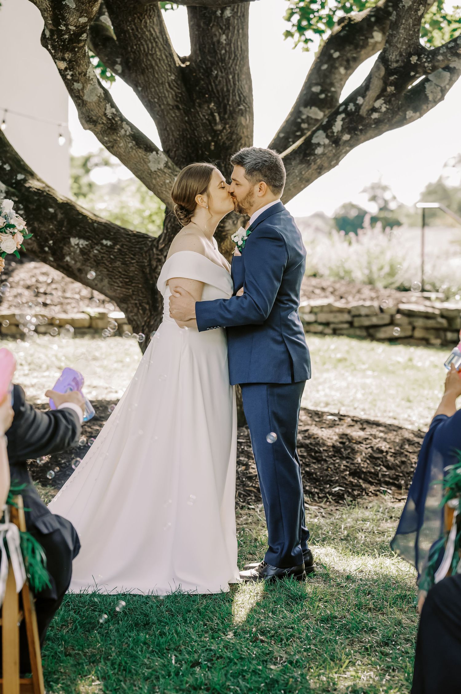 Romantic wedding kiss beneath a sprawling tree with soft natural lighting.