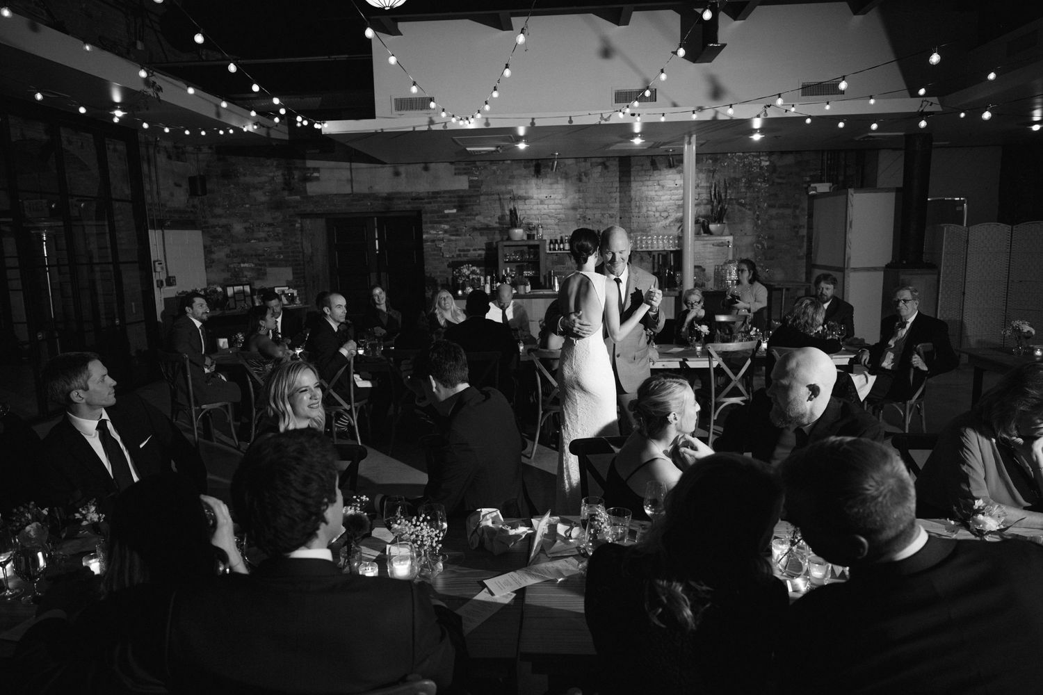 Black and white reception photo showing guests seated around tables with string lights overhead.