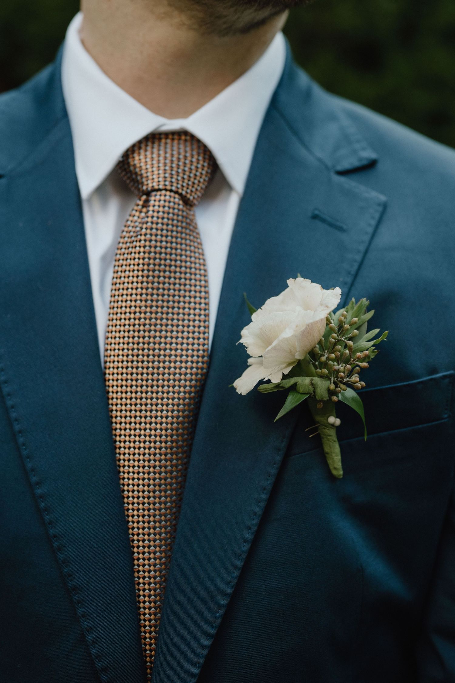 Close-up of a navy blue suit with brown checkered tie and white rose boutonniere.