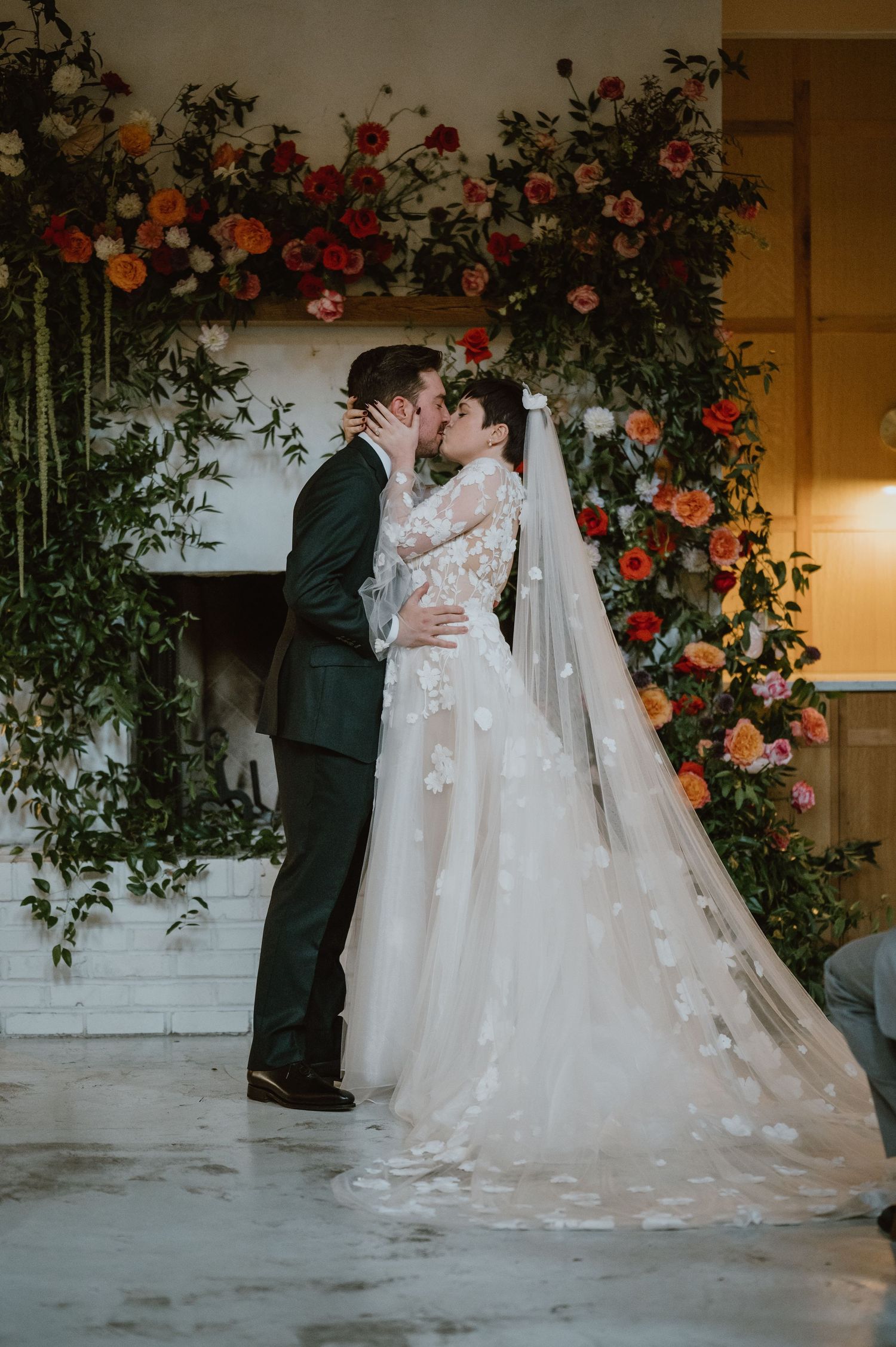 A romantic kiss shared under a floral arch adorned with red and orange blooms at a wedding ceremony.