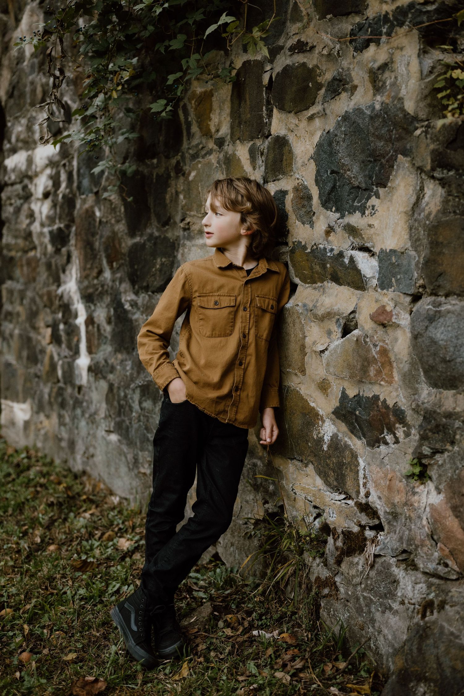 A youth in a brown jacket leans against a rustic stone wall in an autumn setting.