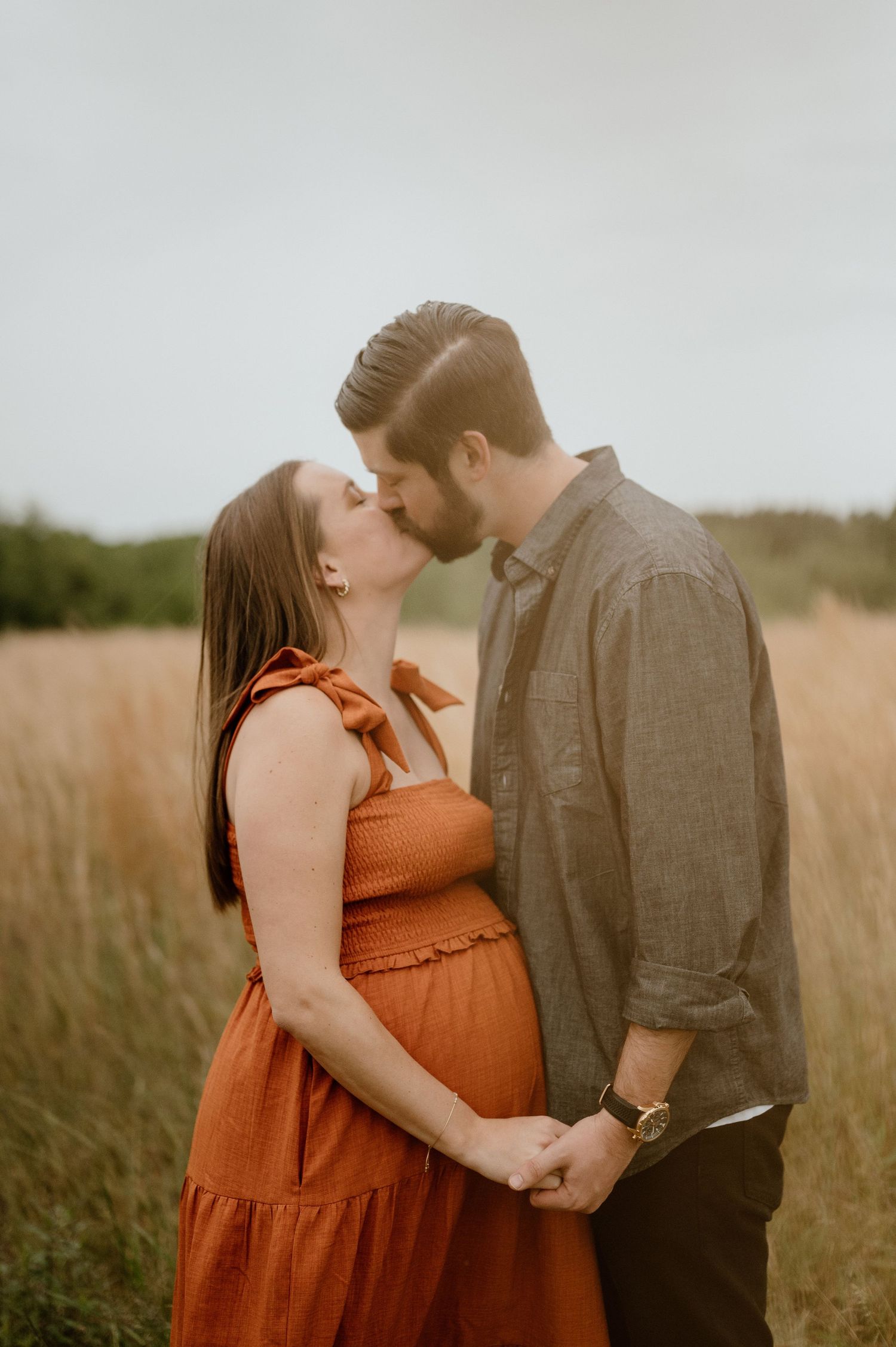 Expecting couple sharing an intimate kiss in a golden grass field at sunset.