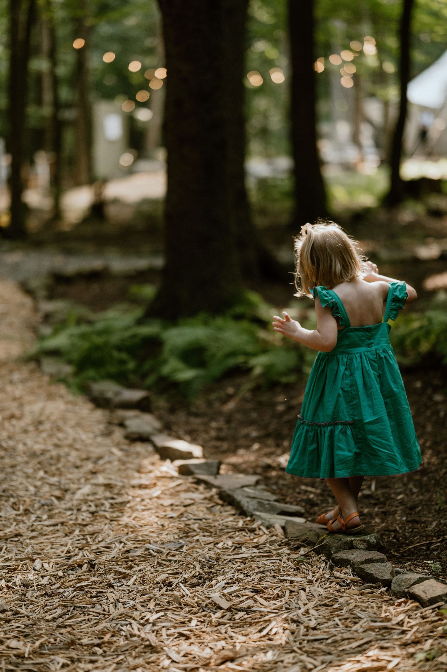 A small child in a green dress dances along a woodland path under string lights.
