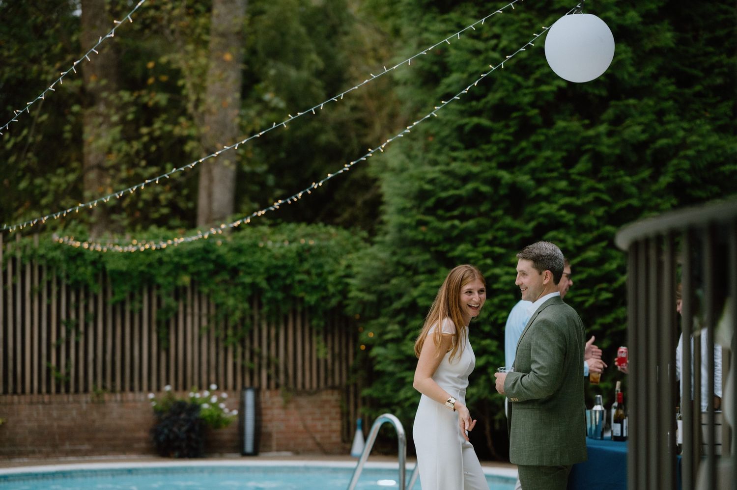 A couple stands near a pool decorated with string lights during an outdoor evening event.