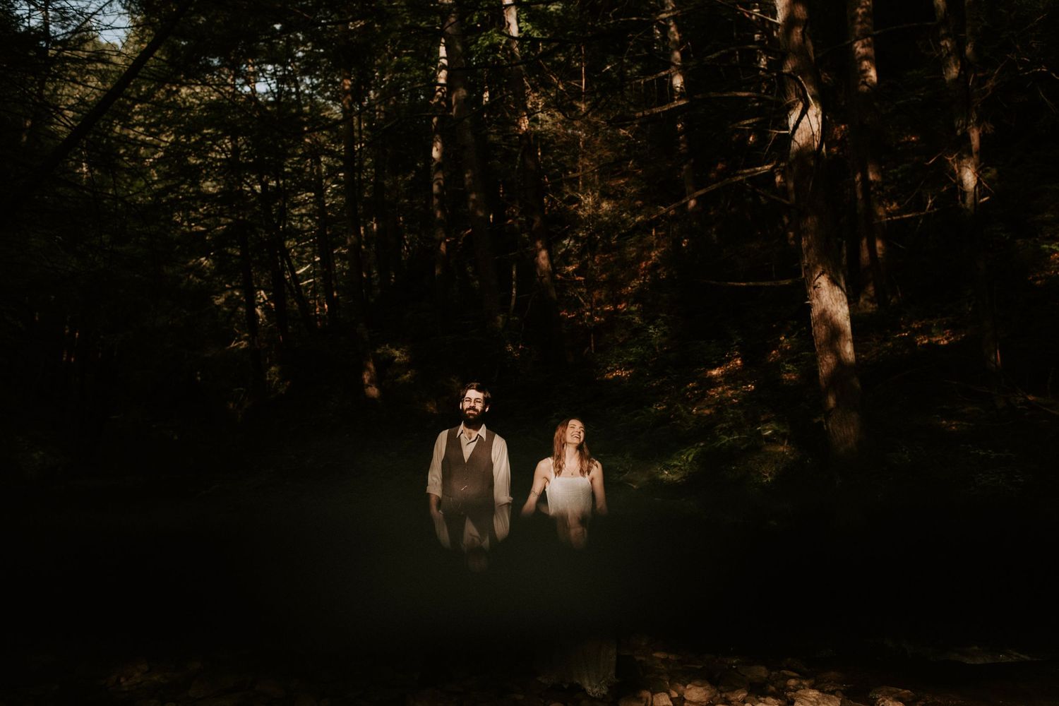 Couple stands together in dark, moody forest with dramatic lighting.