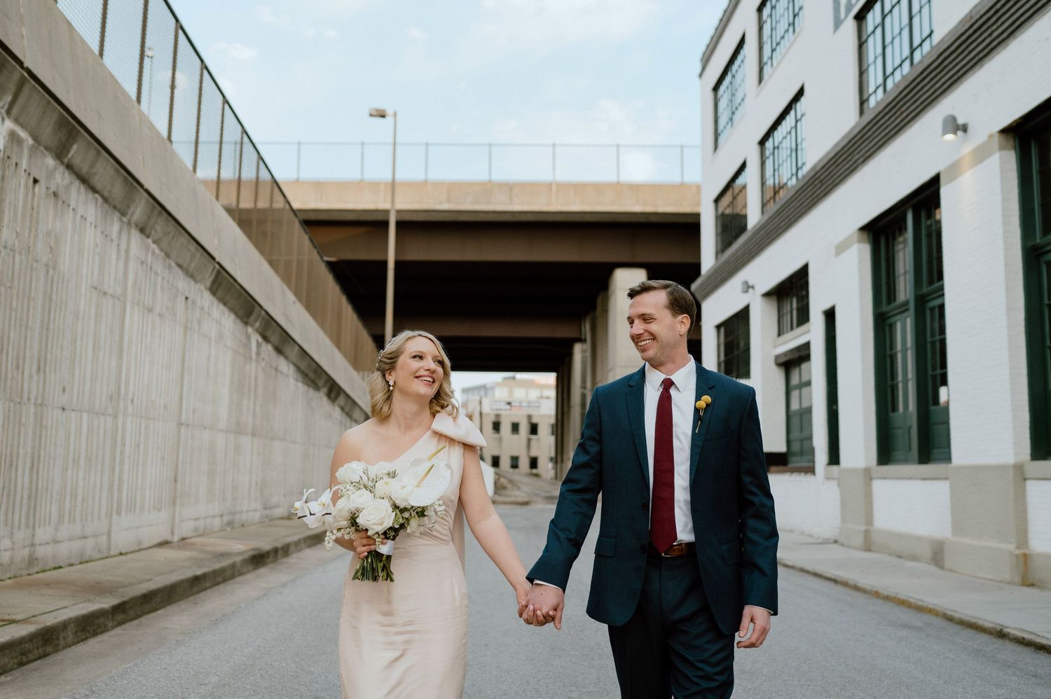 Bride and groom hold hands while walking under an urban overpass.