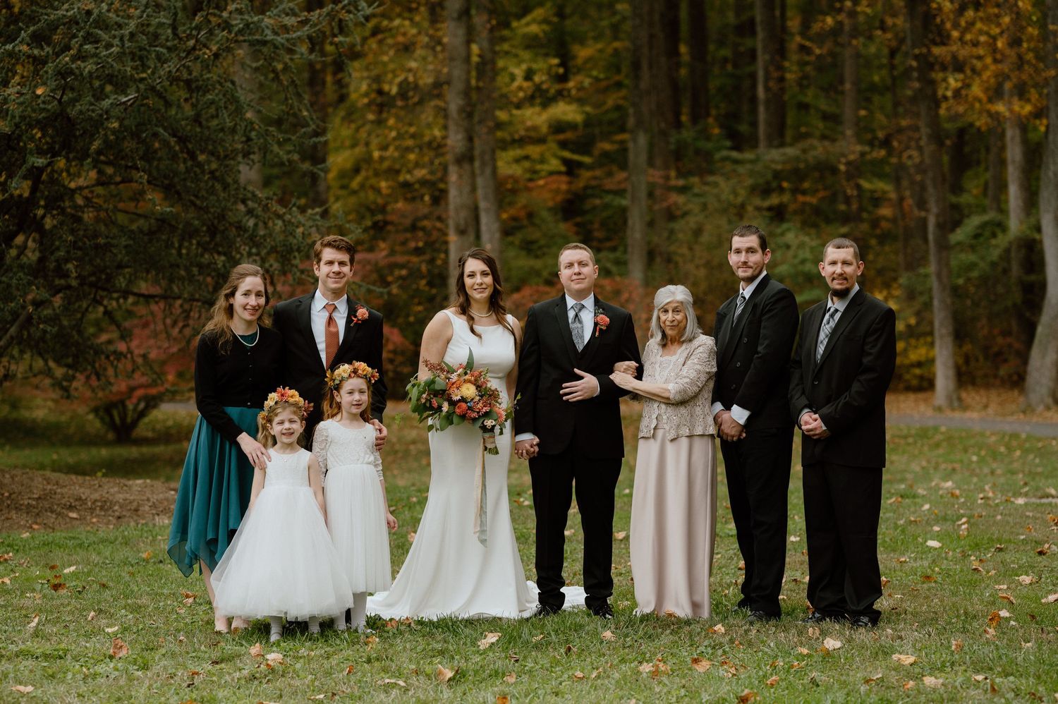 Wedding party gathered outdoors in formal attire against backdrop of autumn trees.
