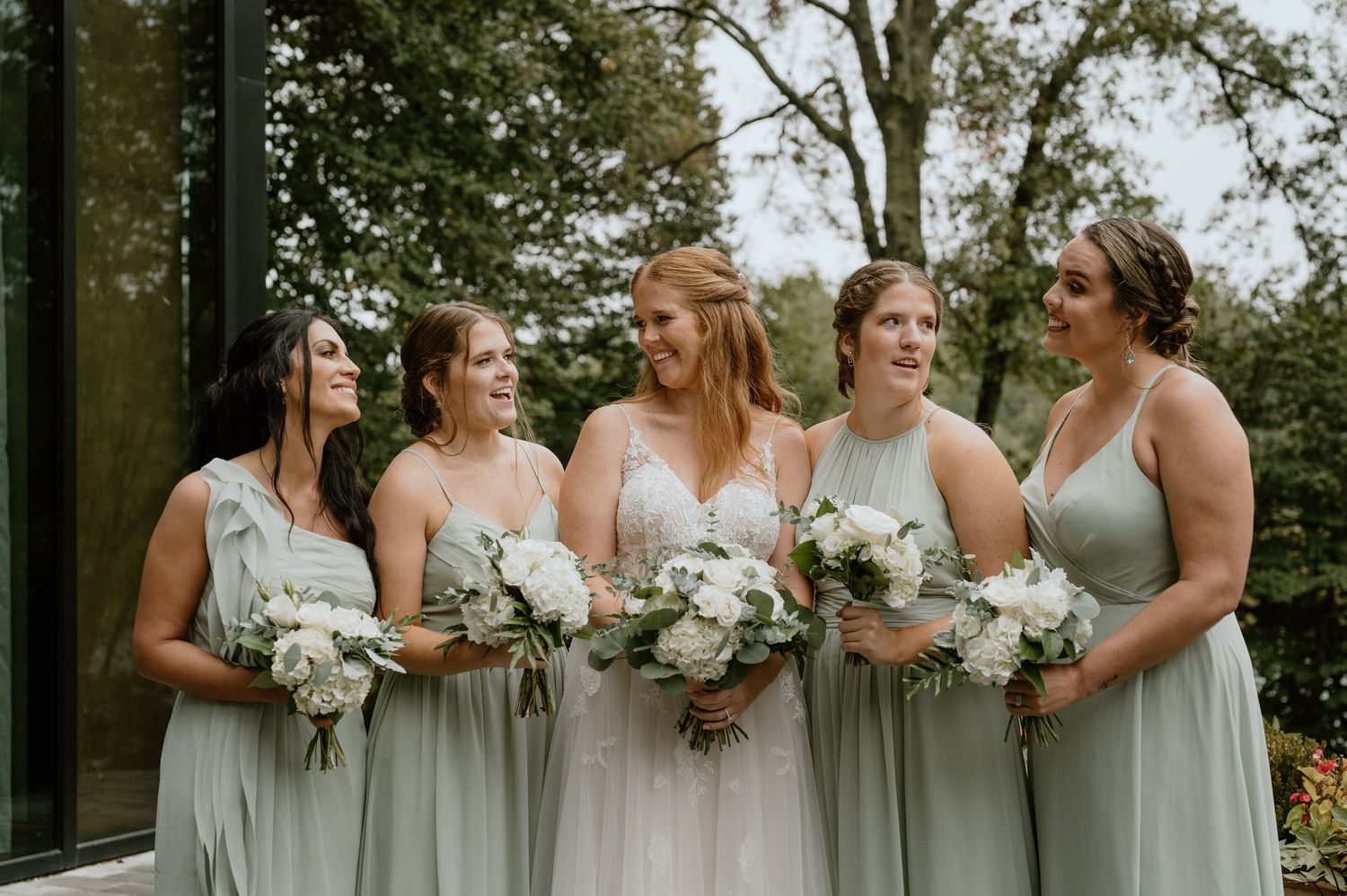 Bridesmaids in sage green dresses hold white floral bouquets while sharing joyful moment outdoors.