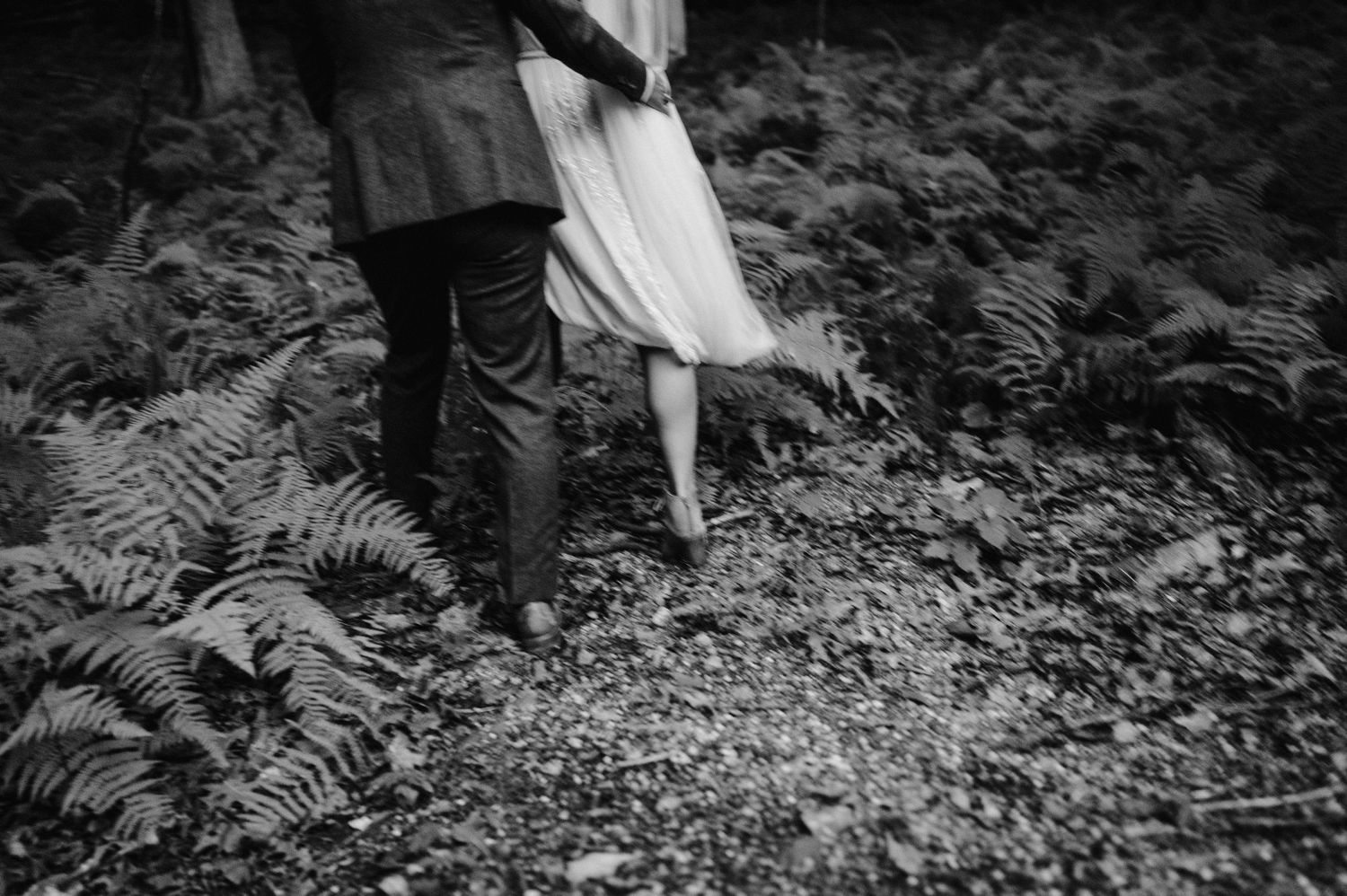 A black and white detail shot of a couple walking through wild ferns in a forest setting.