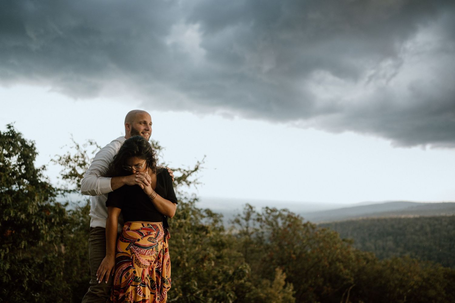 A couple embraces on a mountain overlook with dramatic storm clouds rolling in overhead.