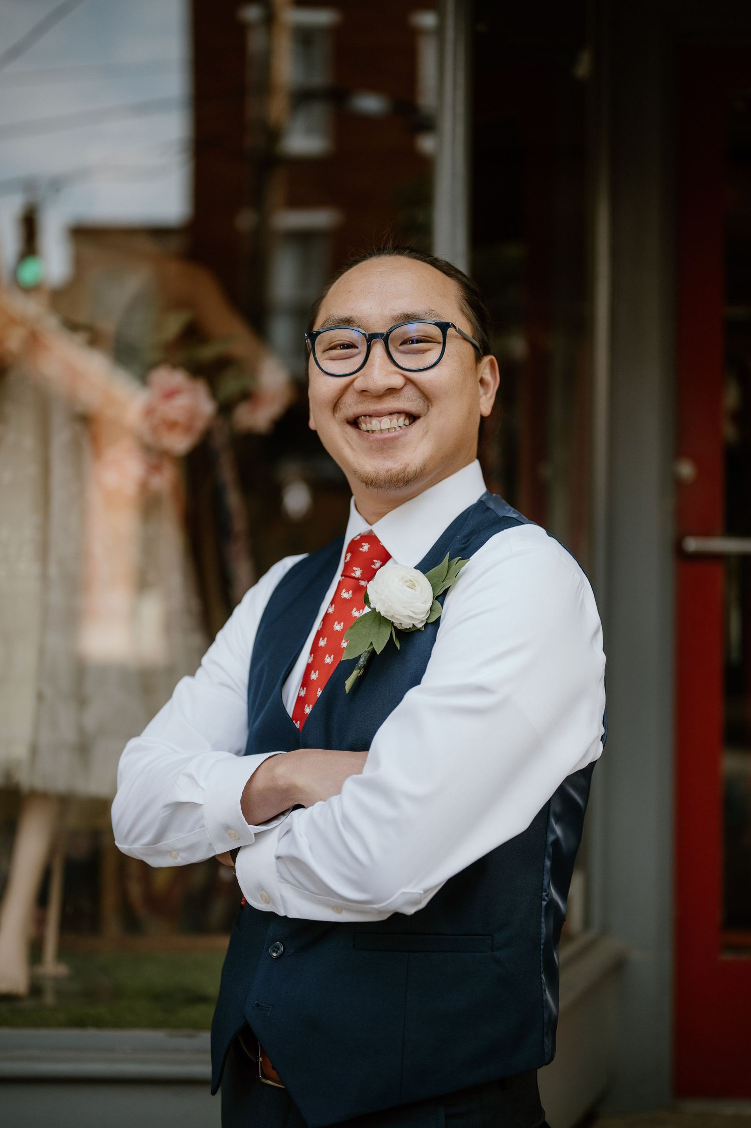 A person in a navy vest and red tie smiles warmly while wearing a white boutonniere.