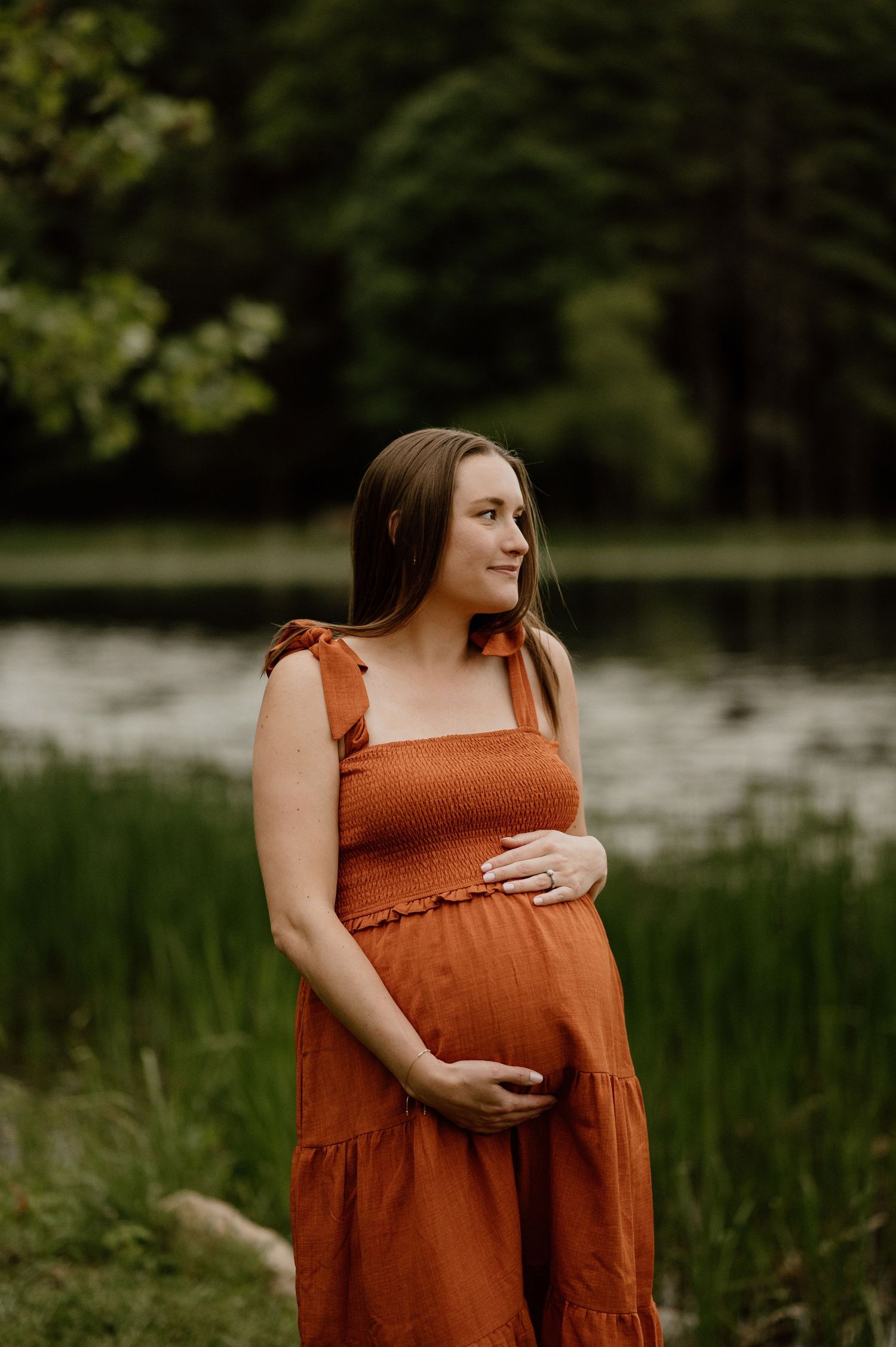 A peaceful maternity portrait by a lake featuring a rust-colored dress with bow details.
