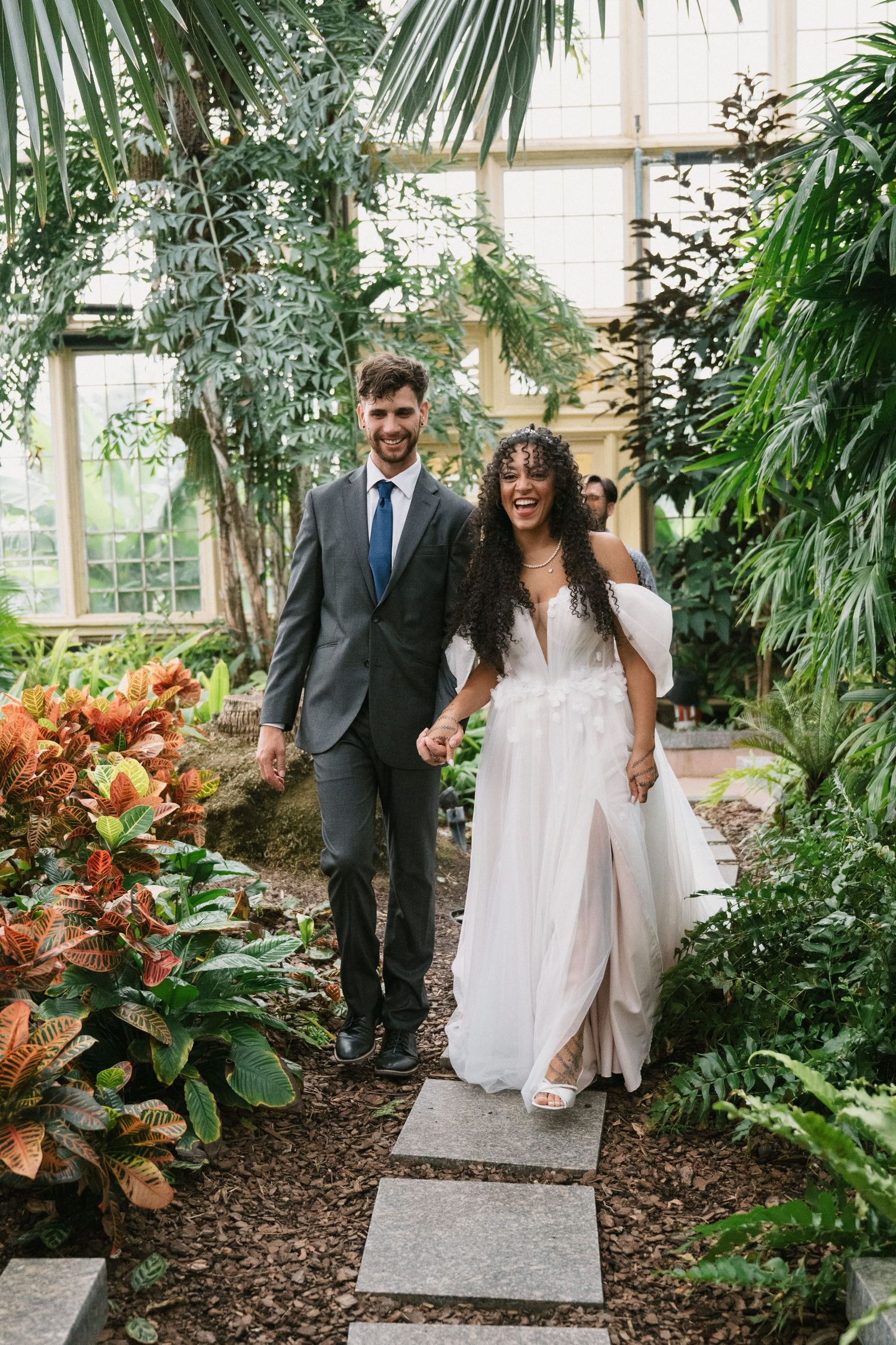 Wedding couple walking through a lush greenhouse filled with tropical plants and orange flowers.