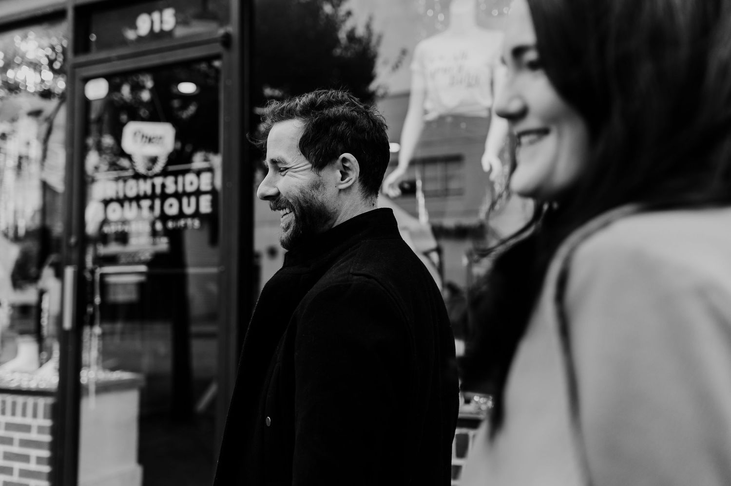 A man in a black coat laughs joyfully outside a boutique shop in a candid black and white street photo.