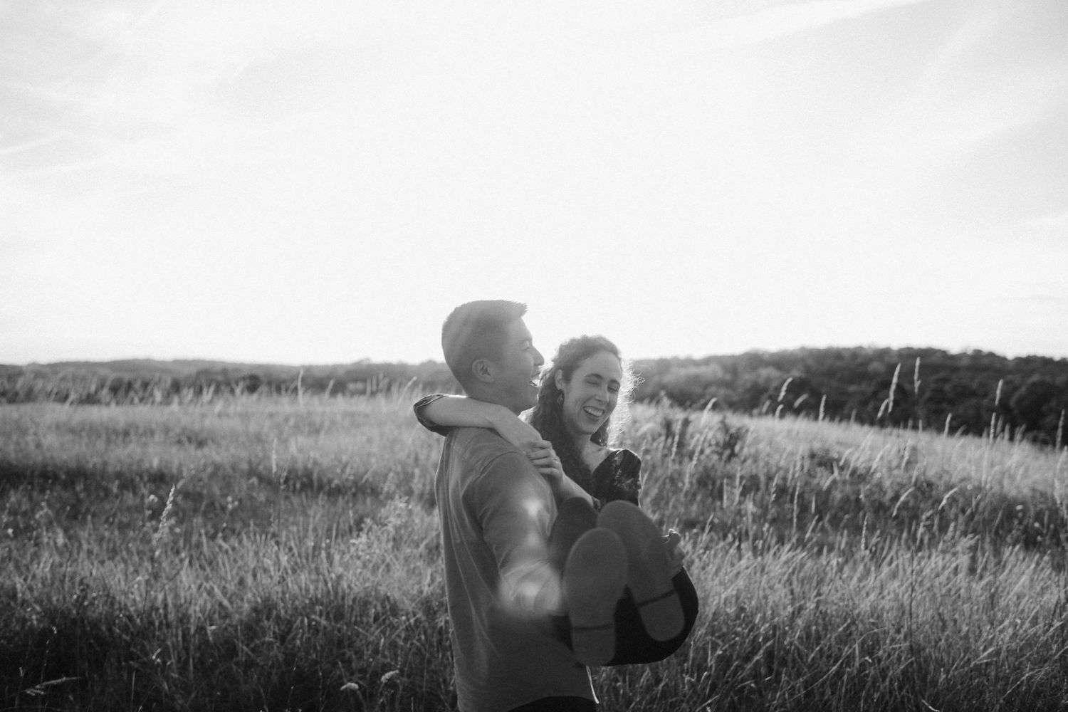 Black and white photo of a couple embracing in a sunlit field with tall grass and trees in the background.