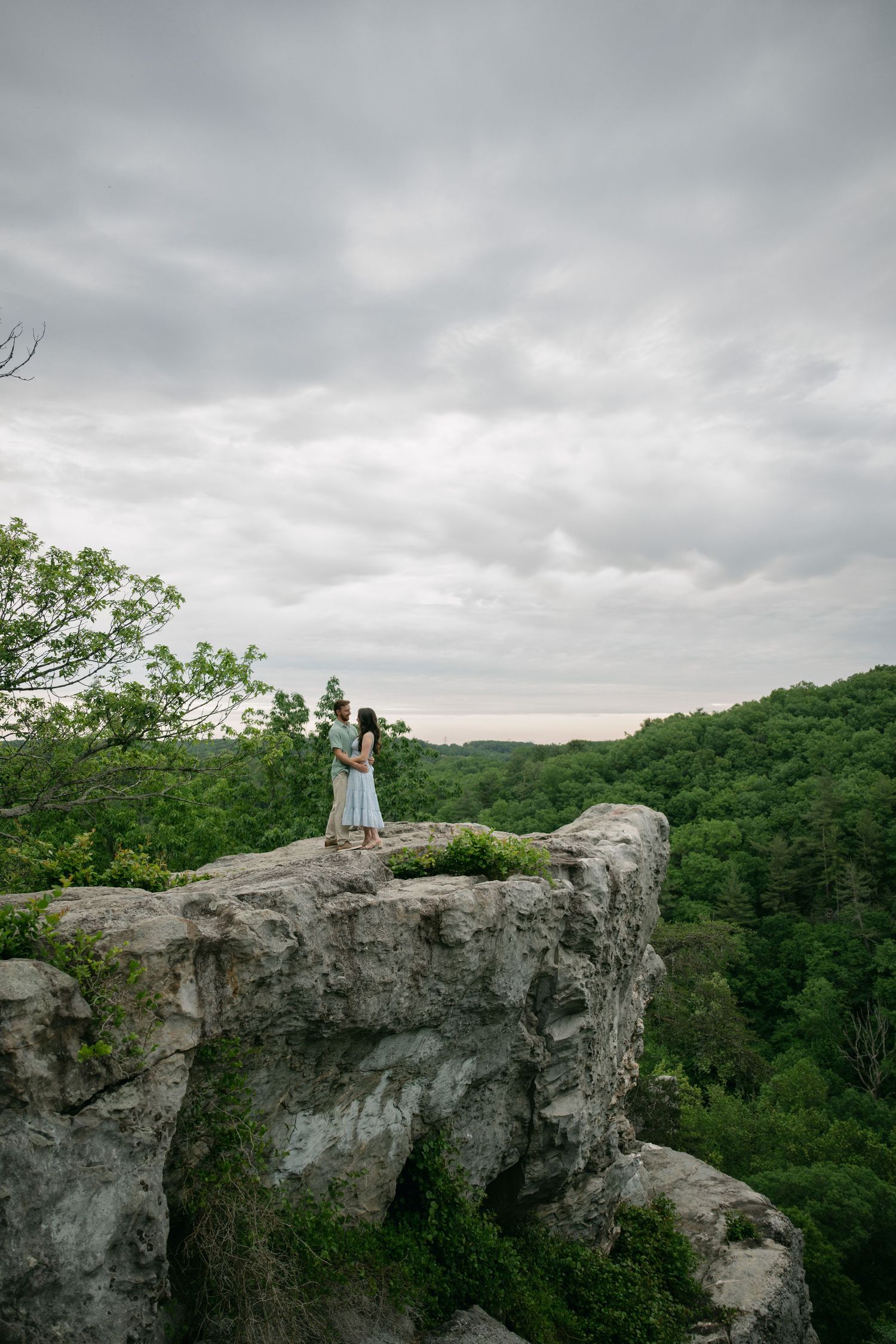 A dramatic rocky overlook provides sweeping views of a forested valley under cloudy skies.