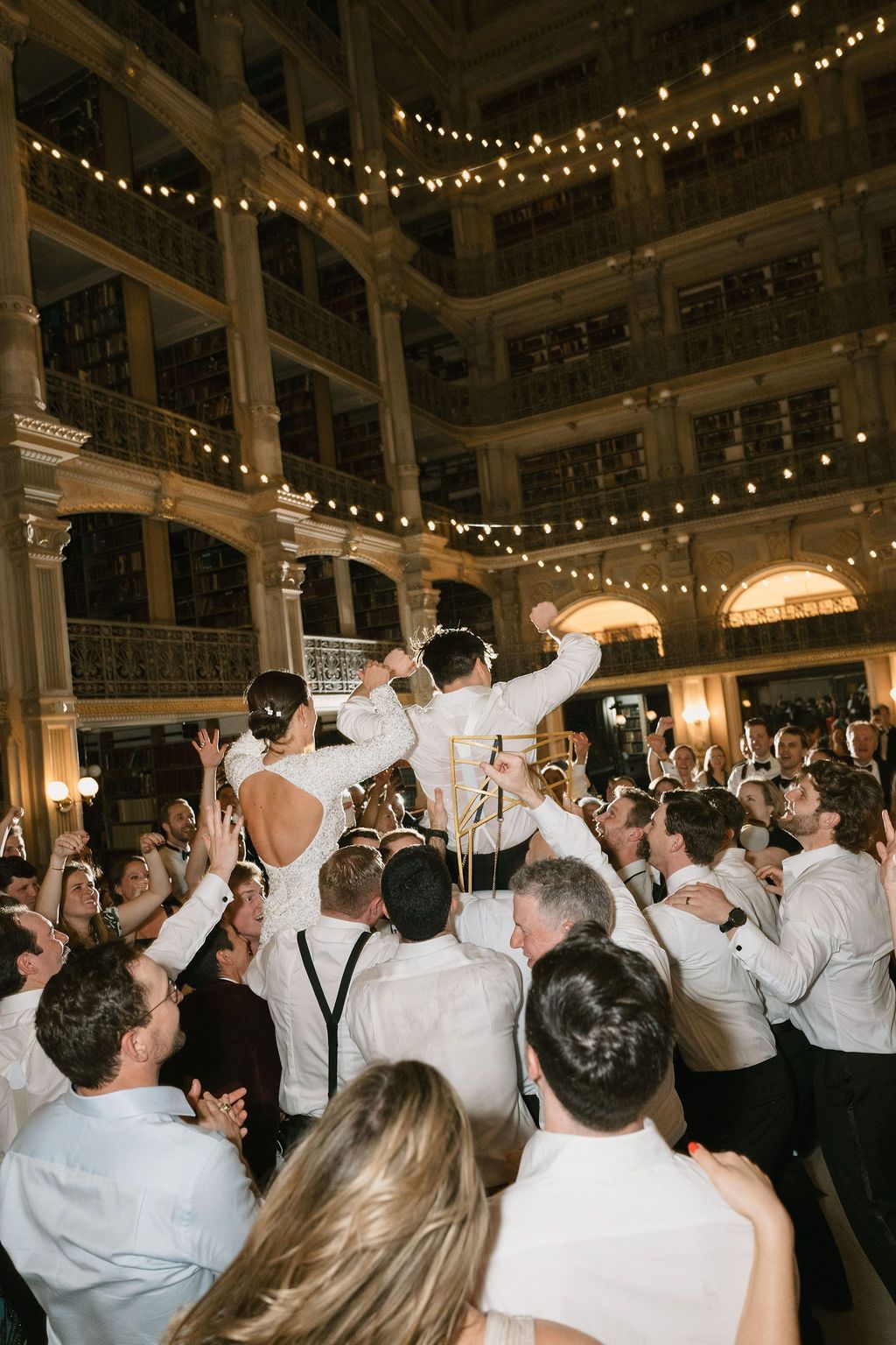 Large crowd celebrating at an indoor wedding reception with string lights overhead at George Peabody Library Wedding in Baltimore