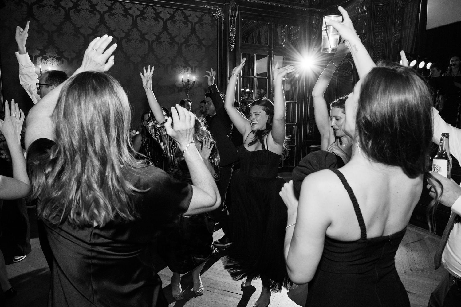 Black and white photo of wedding guests dancing with raised hands on a dark dance floor.