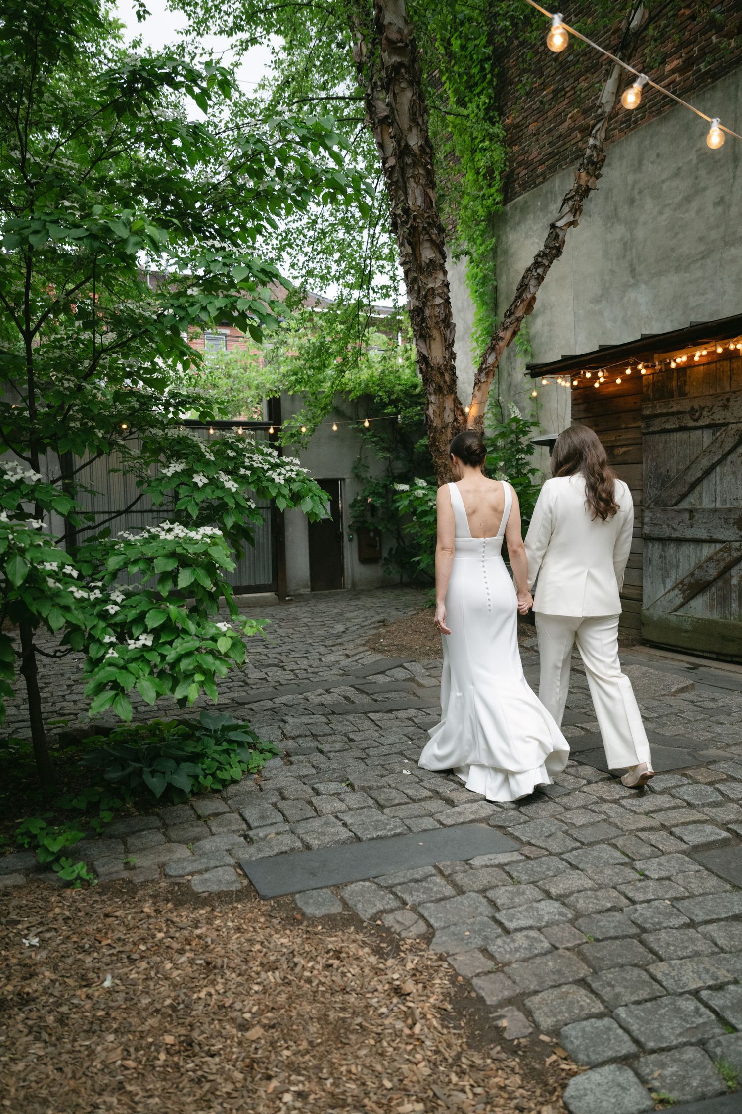 Two people in white dresses walk along a cobblestone path under string lights in a garden setting.