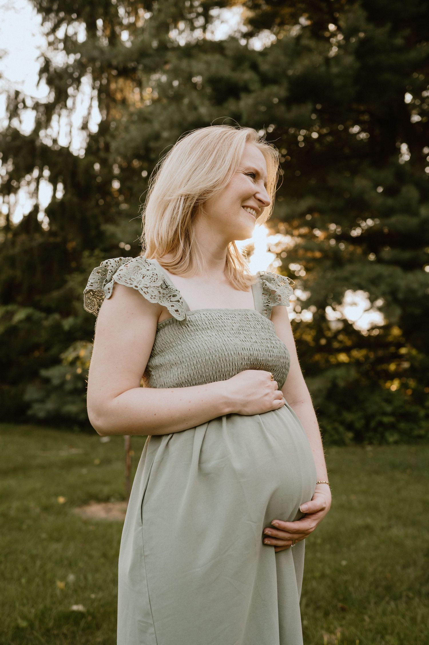 A joyful portrait in a sage green dress against a backdrop of evergreen trees at sunset.