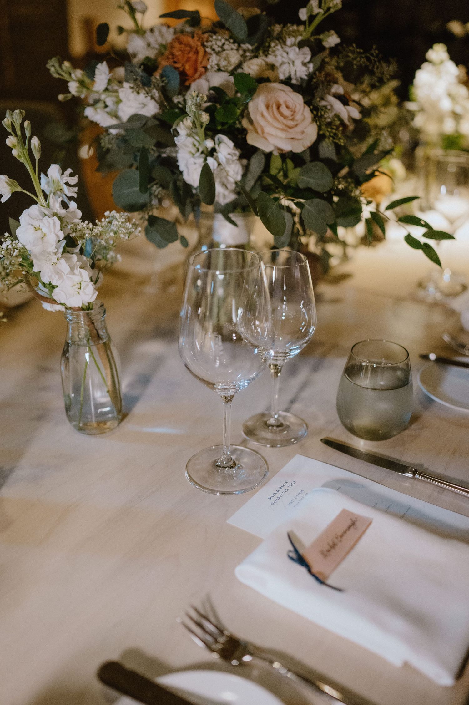 Wedding table setting with blush roses and white floral centerpiece next to wine glasses.
