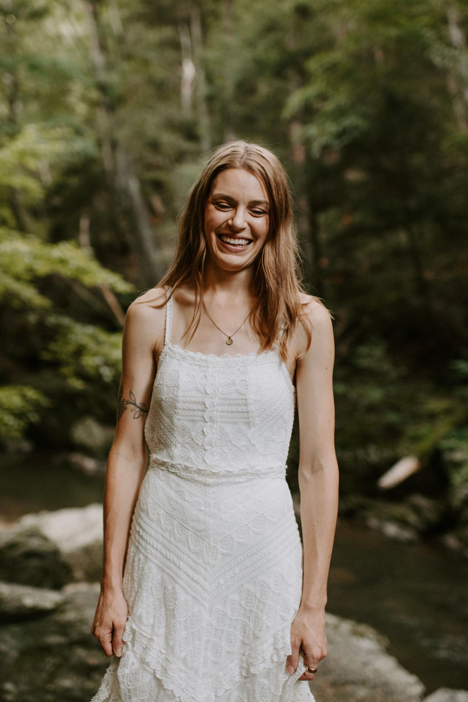 A joyful bride in a white lace dress smiles while standing in a lush forest setting.