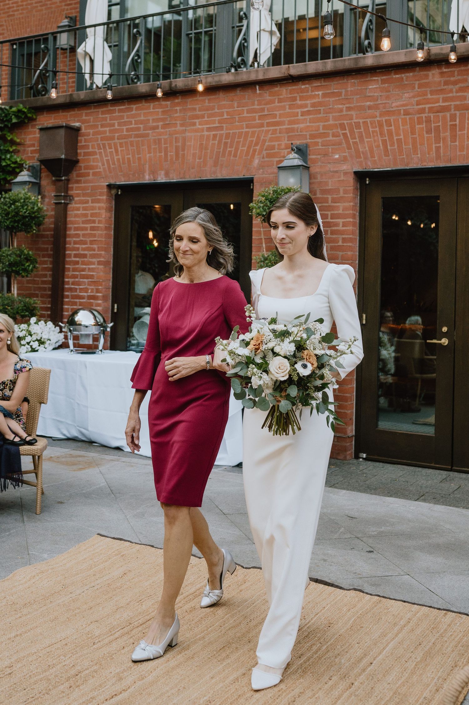 Two elegantly dressed women walking together at an outdoor event, one carrying a floral bouquet.