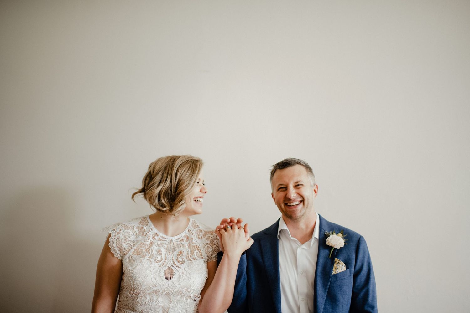 A joyful couple shares a candid moment in formal wedding attire against a neutral backdrop.