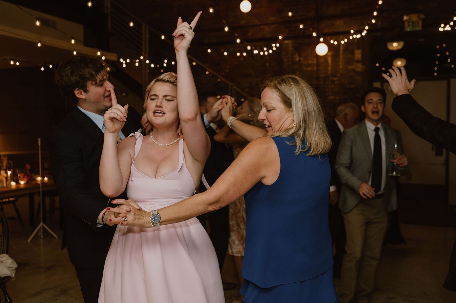 Wedding reception dancers enjoying a lively moment under string lights in a rustic venue.