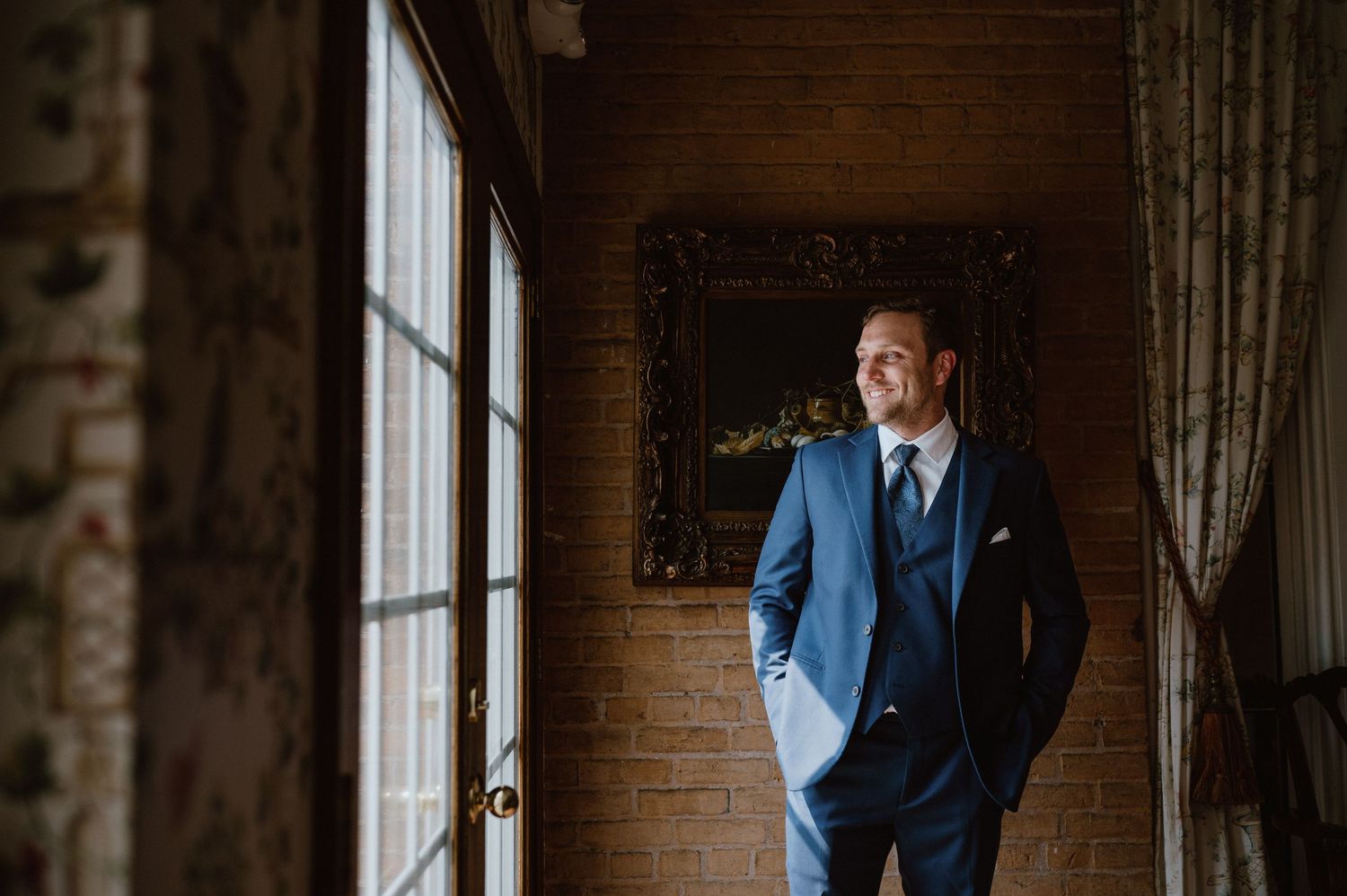 Man in blue suit standing by window in rustic wood-paneled room.
