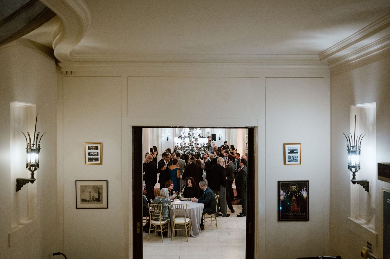 View through doorway of wedding reception with guests gathered in an elegant white-walled room with sconces.