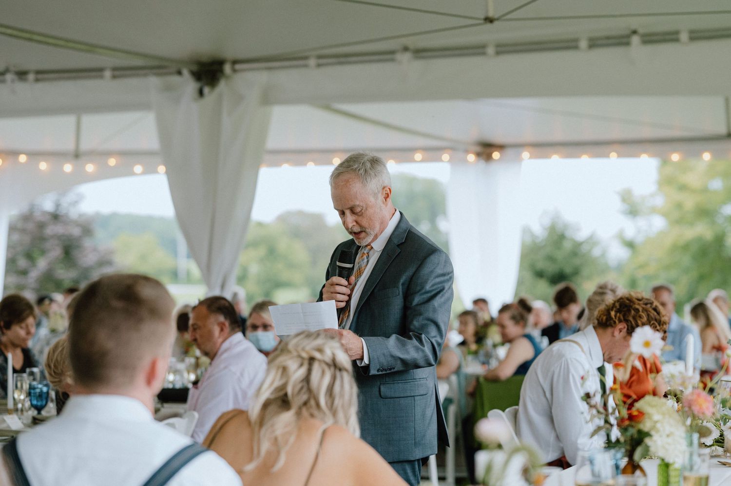 A wedding reception speech being delivered under a white tent with guests seated at decorated tables.
