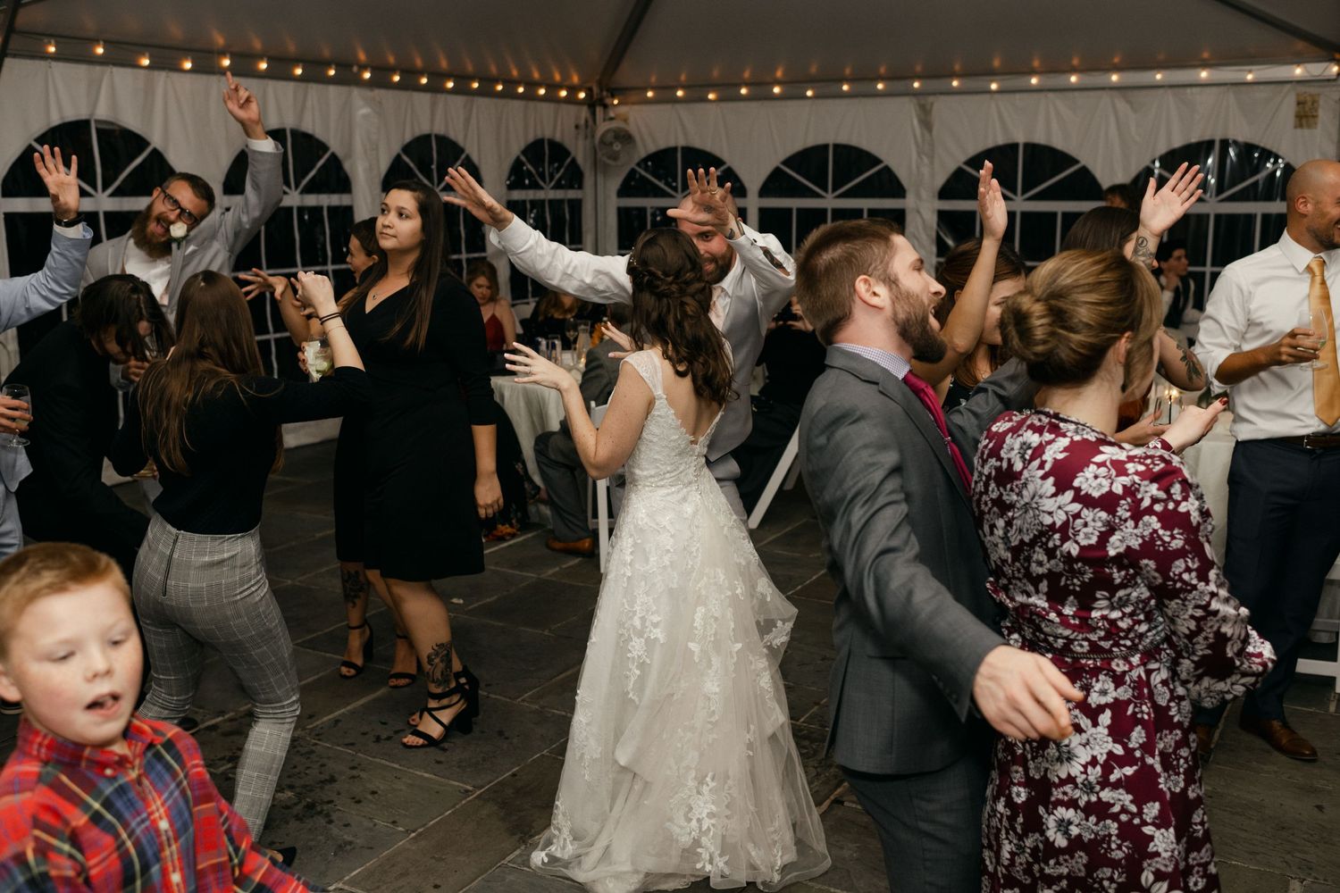 Wedding party and guests dancing energetically under a tent with string lights.