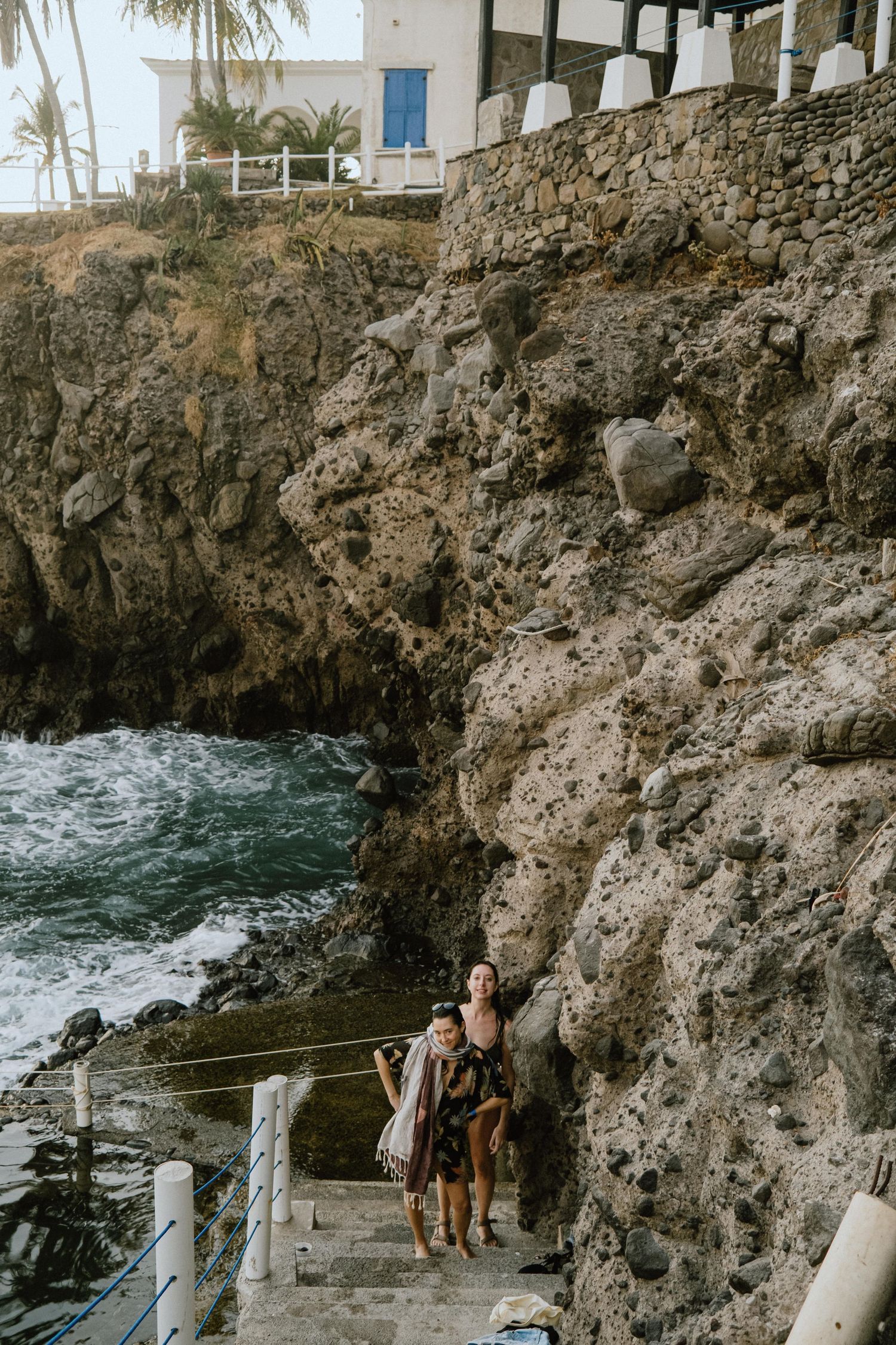 Stone stairway carved into coastal cliffs leads down to ocean pool with swimmers.