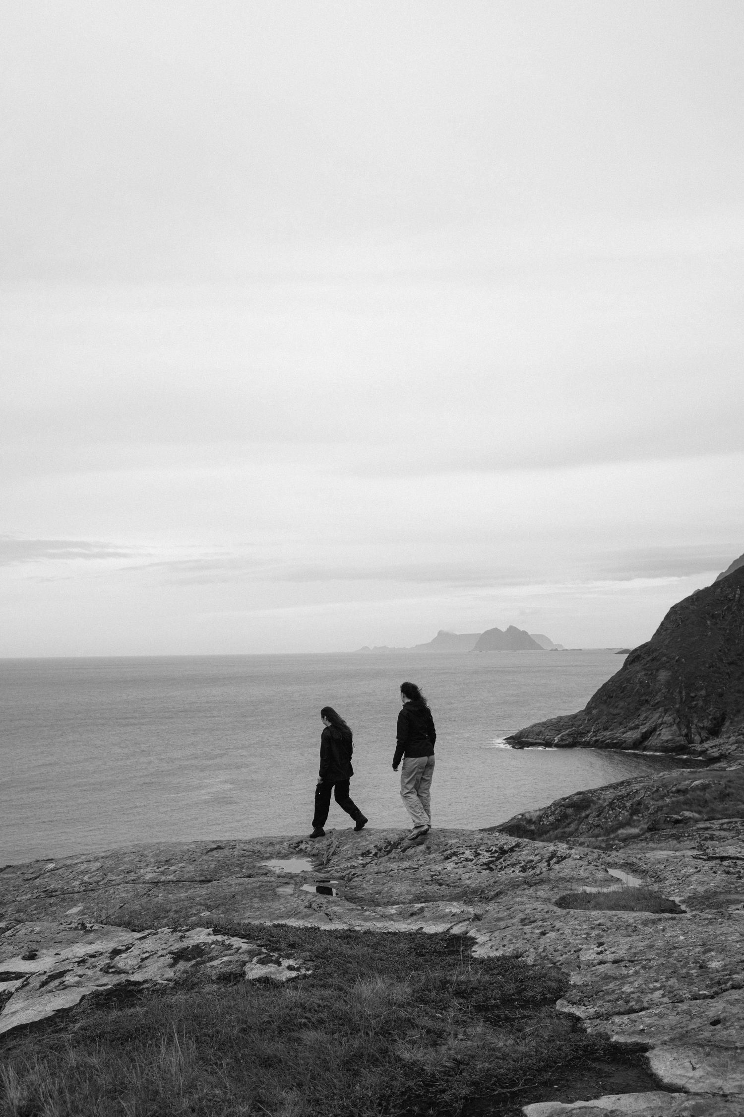 Silhouettes on coastal cliff edge overlooking distant islands in black and white.