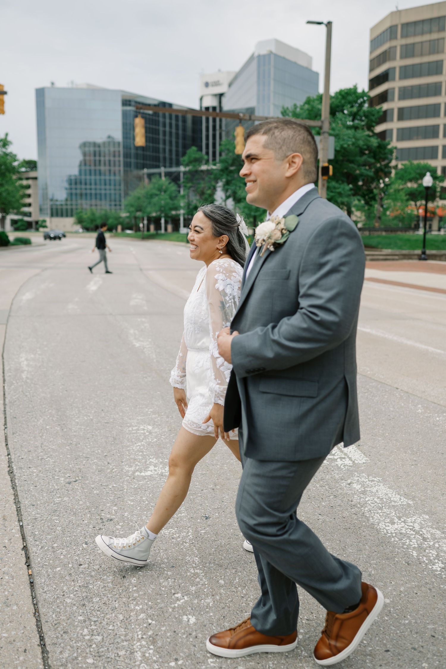 A couple in wedding attire walks together on a city street with modern buildings in the background.