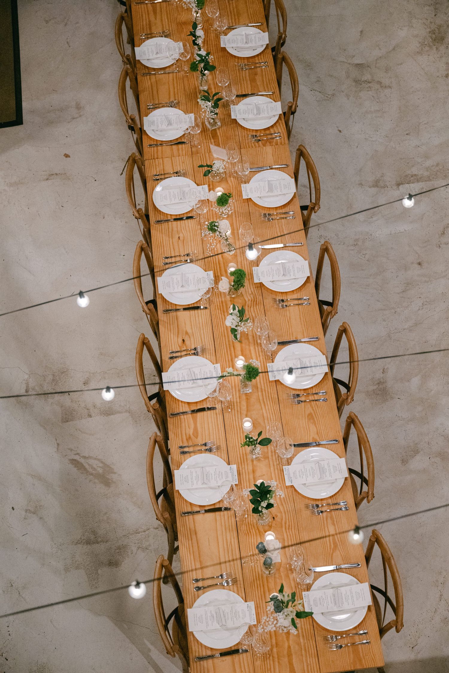 Overhead view of an elegant wedding table setting with wooden chairs and white place settings decorated with greenery.