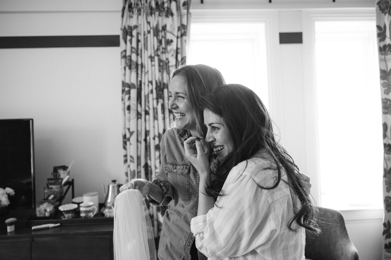 Two women share joyful laughter during wedding preparations in a bright room.