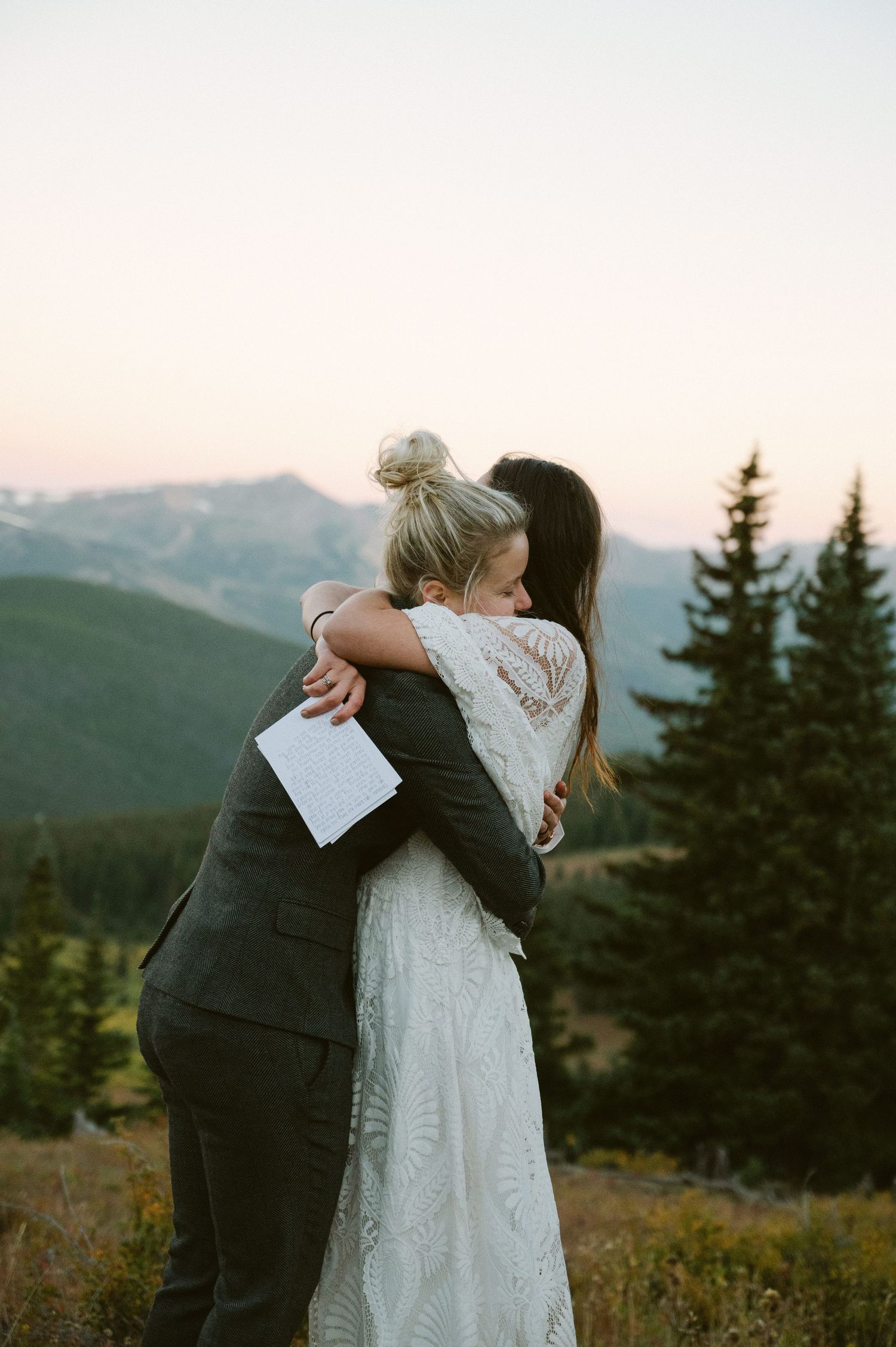 Two people embracing in white and dark attire against mountain landscape at dusk.