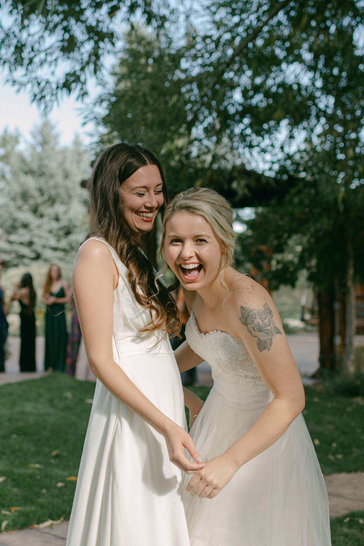 Two women in white dresses laughing joyfully together at an outdoor wedding.