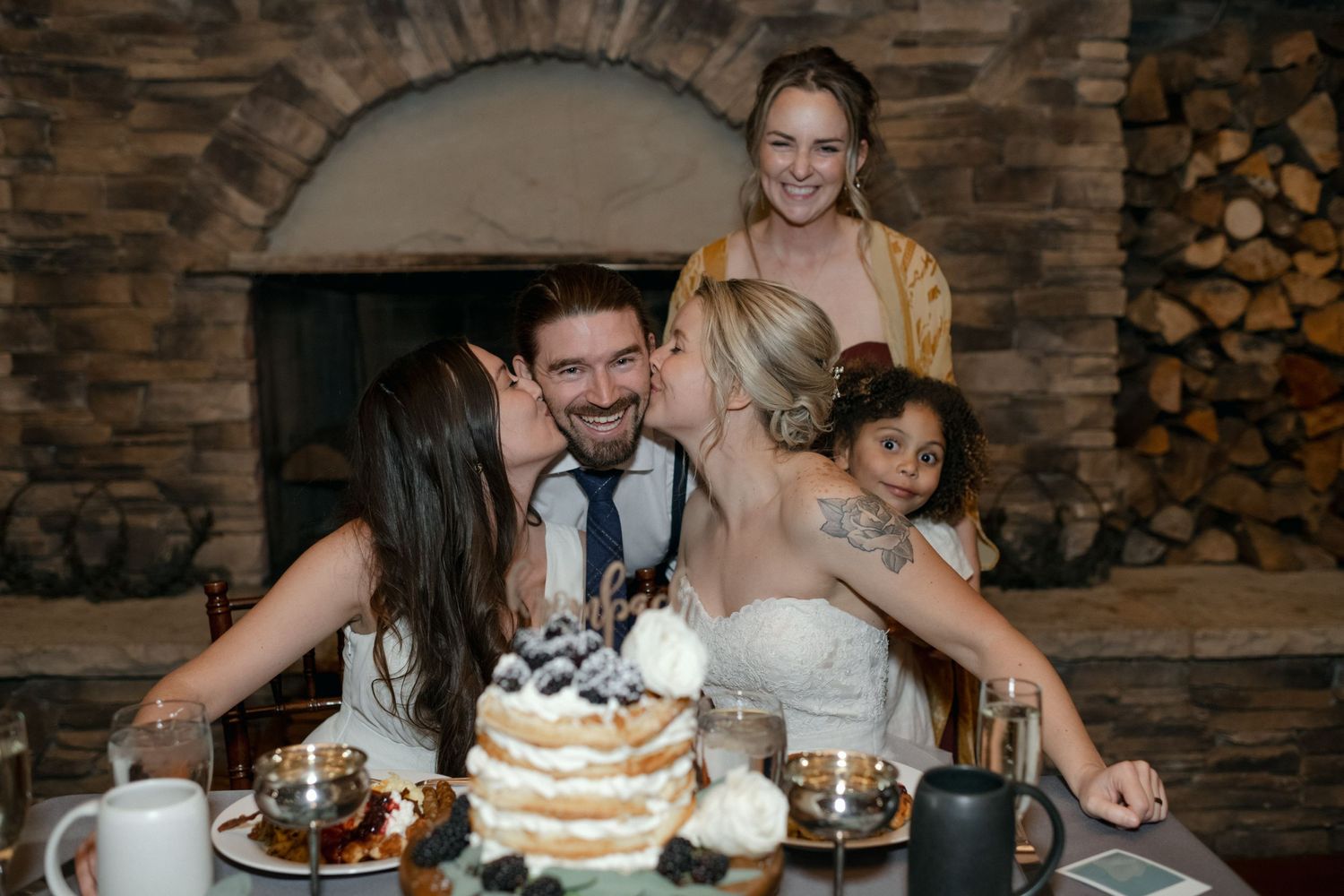 Wedding party gathers around a rustic cake table with stone fireplace backdrop.