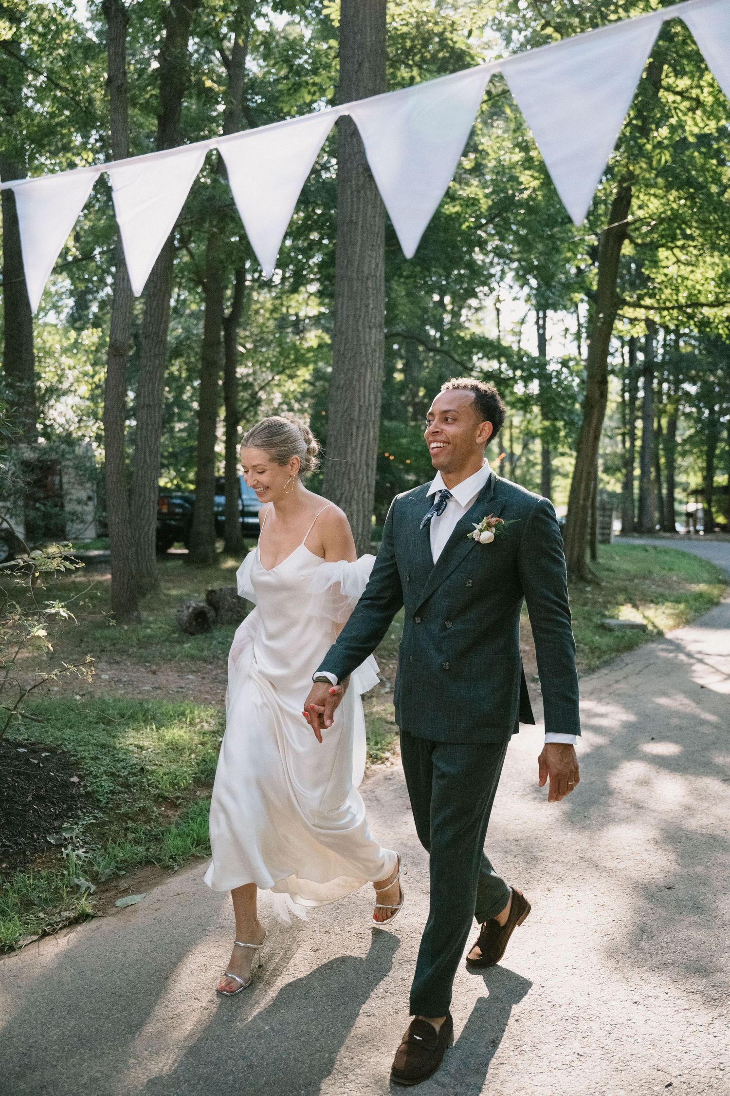A newlywed couple walks beneath white triangular bunting decorations on a wooded path.