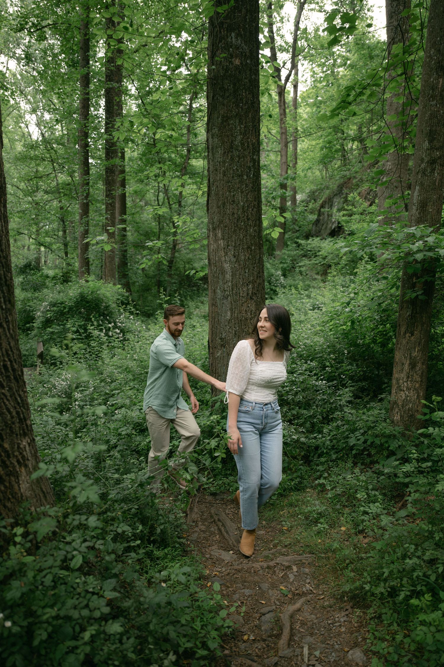 Couple holding hands walking through a lush green forest trail surrounded by tall trees.
