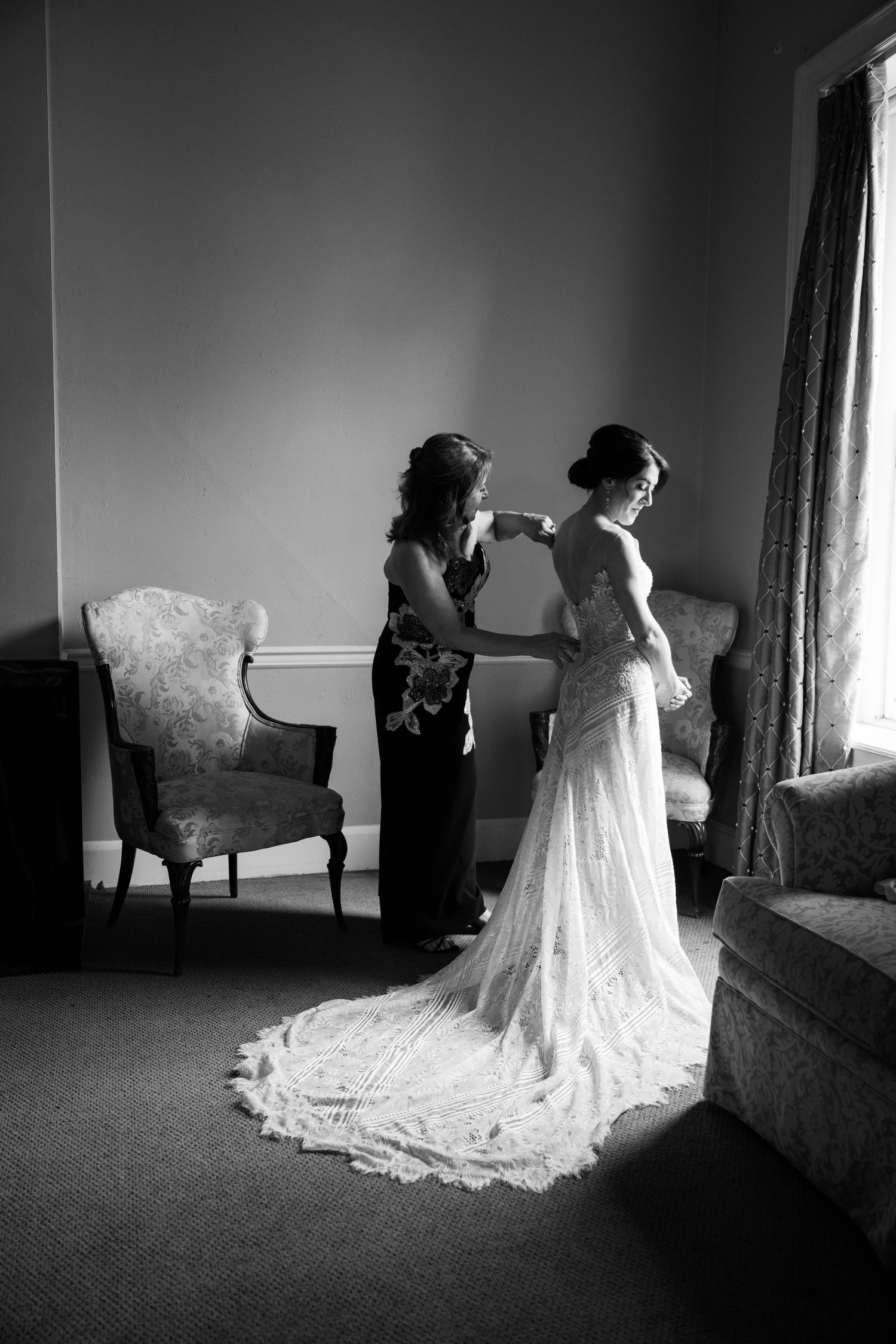 Black and white photo of a wedding dress being adjusted in a room with vintage furniture.