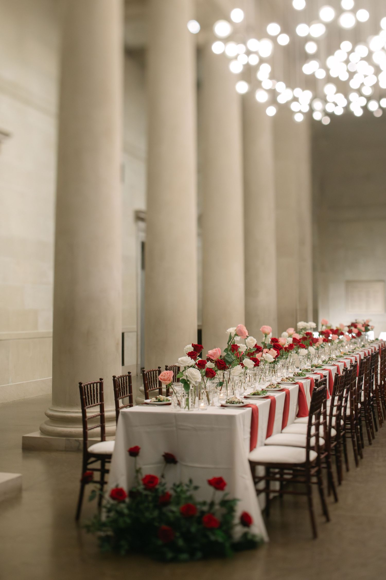 Elegant wedding reception table with red roses and hanging lights in columned hall.